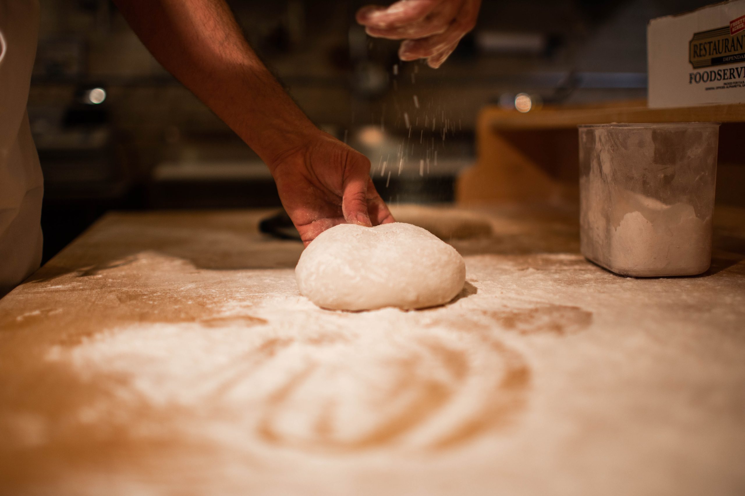 A hand sprinkling flour over a round piece of dough on a wooden surface. - International Hotel School