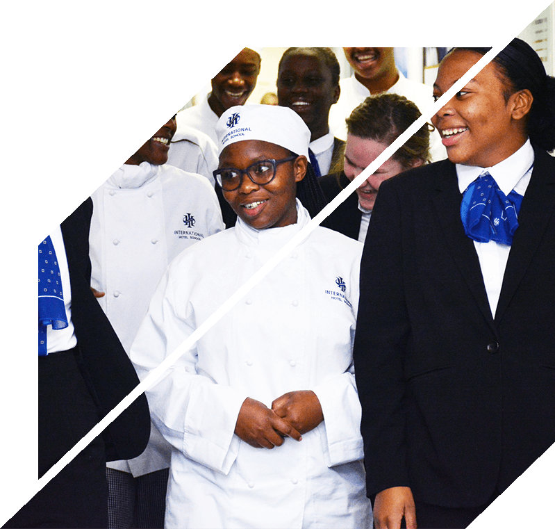 Group of culinary students wearing chef uniforms and formal attire, smiling and interacting in a kitchen setting.