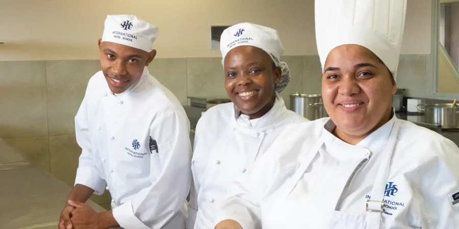 Three culinary students and an instructor wearing white chef uniforms in a professional kitchen setting. - International Hotel School